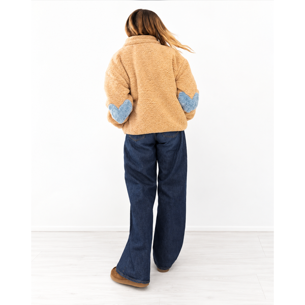 A woman is shown from behind wearing a tan sherpa fleece jacket with blue heart-shaped elbow patches and dark blue jeans, standing against a plain white wall.