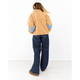 A woman is shown from behind wearing a tan sherpa fleece jacket with blue heart-shaped elbow patches and dark blue jeans, standing against a plain white wall.
