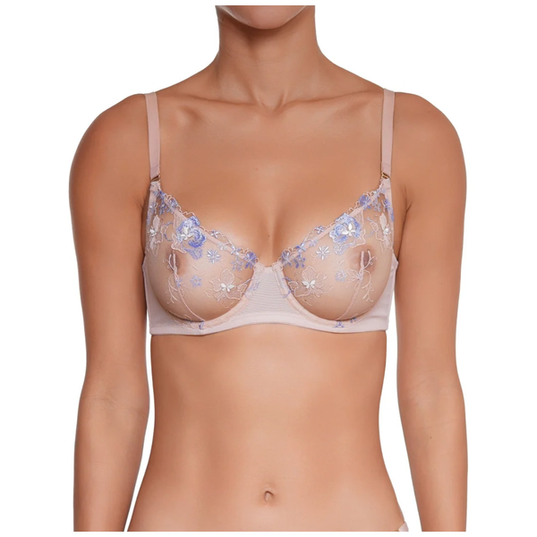 A woman wearing a light beige underwire bra with blue floral embroidery and sheer mesh cups, shown from the neck to the lower torso against a plain white background.