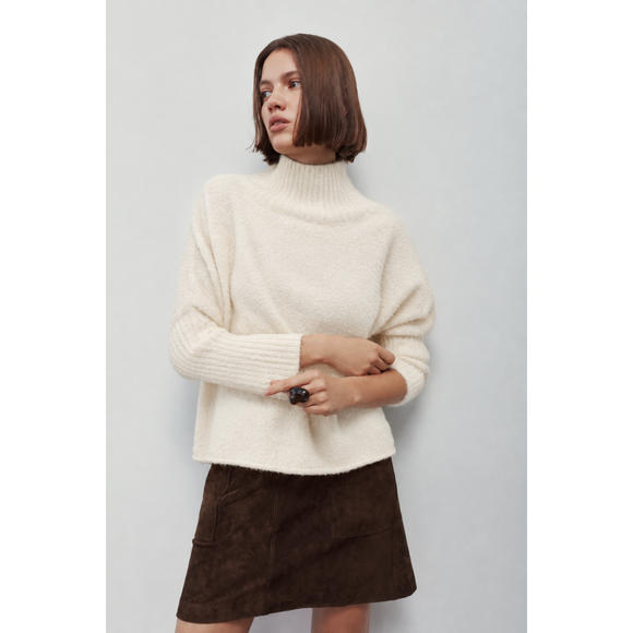 A woman wearing a cream-colored, fuzzy ribbed turtleneck sweater and a dark brown mini skirt, standing against a plain light gray background.