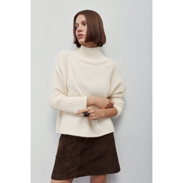 A woman wearing a cream-colored, fuzzy ribbed turtleneck sweater and a dark brown mini skirt, standing against a plain light gray background.