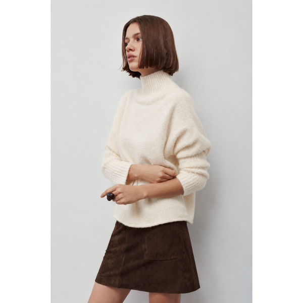 A woman wearing a fuzzy off-white mock-neck knit sweater and a brown corduroy mini skirt, standing against a light gray studio background.