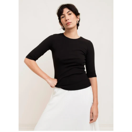 A model wearing a black 3/4-sleeve crewneck top and white skirt, posing against a plain off-white studio background.