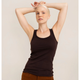 A woman with a shaved head wearing a dark brown ribbed tank top and brown pants, posing with her arms raised against a light beige studio background.
