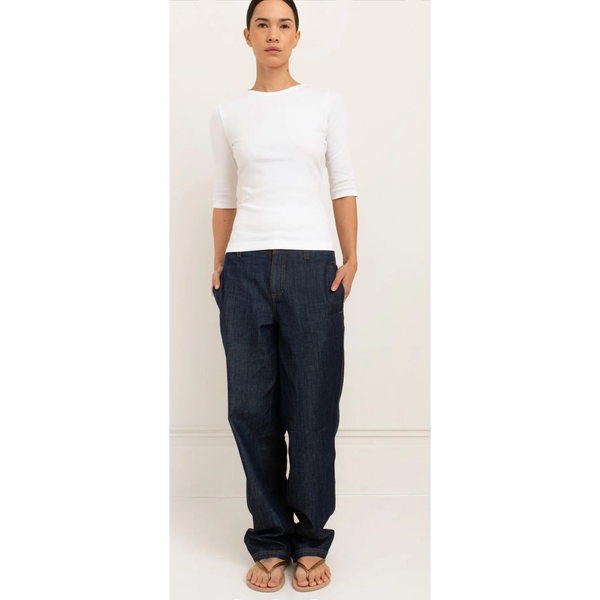 A woman modeling dark blue wide-leg denim jeans with a white three-quarter sleeve top and tan sandals, standing against a plain off-white wall.