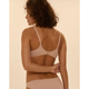A woman with curly brown hair is shown from the back wearing a light beige underwire bra and matching underwear against a solid light beige background.