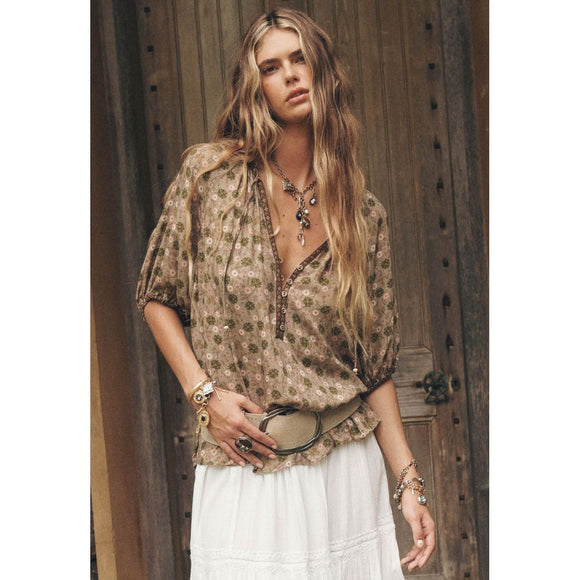 Woman wearing a patterned blouse and white pants standing in front of a wooden door.