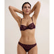 A woman modeling a matching lace lingerie set in a deep plum purple color, featuring a sheer underwire bra and coordinating high-waisted briefs, set against a plain light beige background.