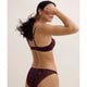 A woman modeling a dark plum lace bra and matching lace panties against a plain light beige studio background.
