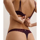 A close-up back view of a model wearing a dark plum lace bralette and matching thong with gold-tone hardware against a light beige studio background.