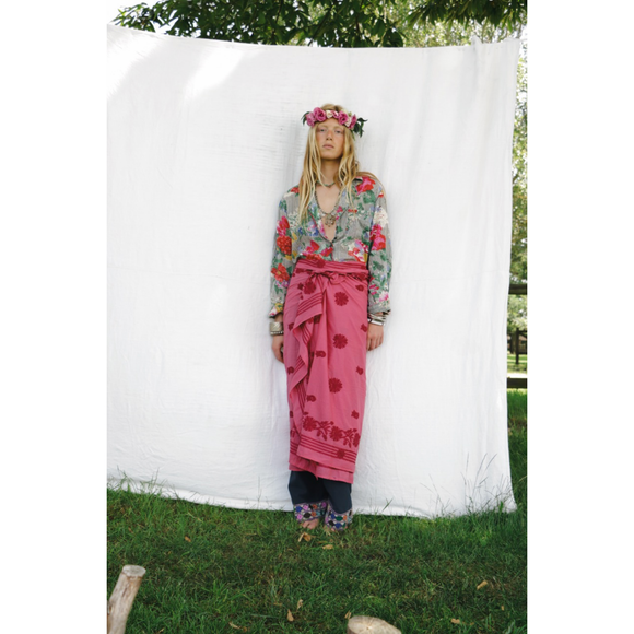 Woman in a floral top and pink sarong skirt standing against a white backdrop outdoors.