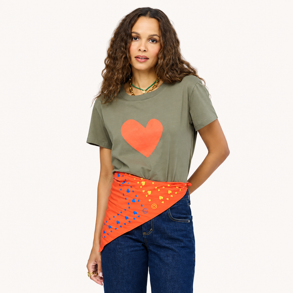 A woman modeling an olive green t-shirt with a large orange heart on the front, paired with blue jeans and an orange patterned bandana tied around her waist, against a plain off-white background.