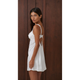 A woman wearing a white satin slip dress with lace trim and an open back, posing against a dark brown wooden paneled wall.