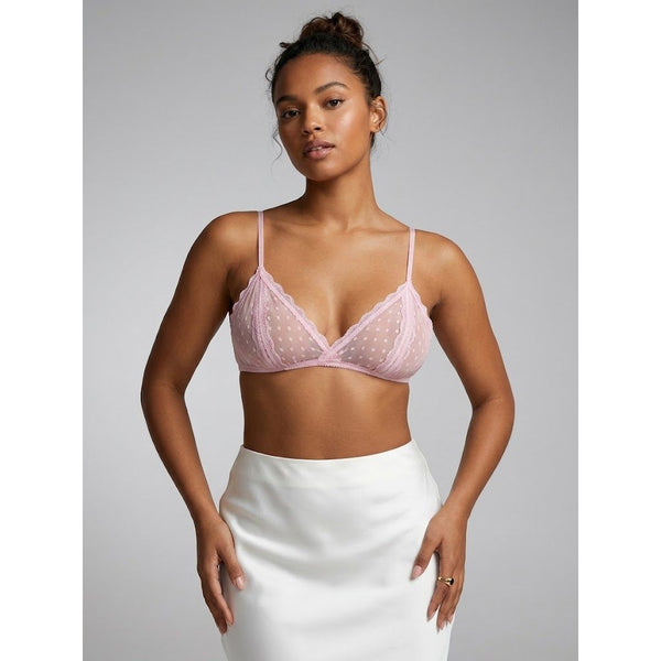 A woman modeling a light pink sheer mesh bralette with scalloped edges and dotted pattern, paired with a white satin skirt, against a solid gray studio background.