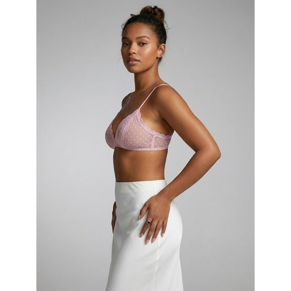 A woman modeling a light pink sheer mesh bralette with white polka dots, paired with a white skirt, against a solid gray studio background.