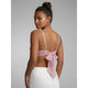 A woman shown from the back wearing a light pink strappy top with a bow detail and white bottoms, photographed against a plain light gray studio background.