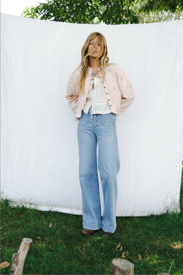Woman in a light jacket, off white button tank and jeans standing against a white wall with greenery.